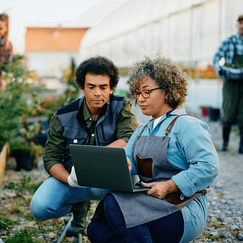 illustration-mission-professionnelle Une femme en tablier et un homme, tous deux à genoux, regardent l'écran d'un ordinateur portable dans un jardin partagé urbain