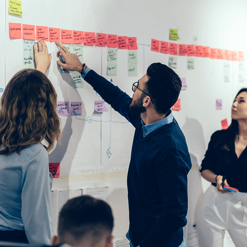 illustration-perspective-eco-econseiller Un homme montre du doigt un tableau blanc plein de post-its devant 2 femmes