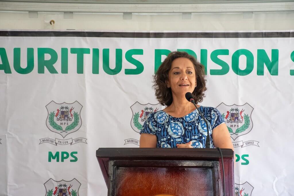 femme a robe à motifs bleus qui parle à un micro sur une scène devant une affiche avec le logo MPS : Mauritius prison service